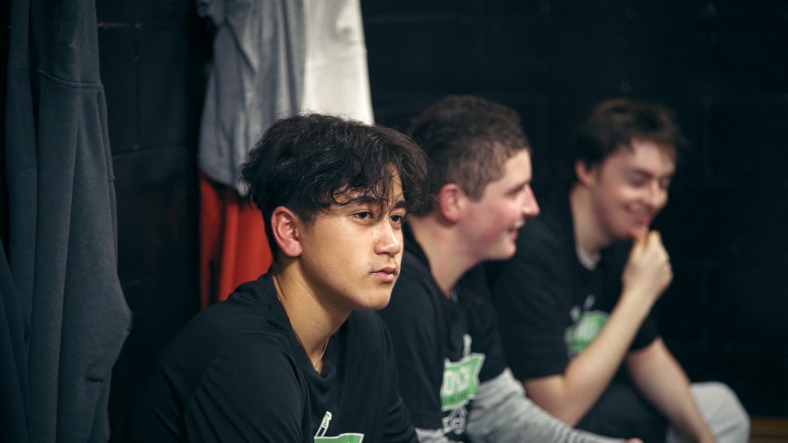 Three young players in Buddy Check shirts in a locker room