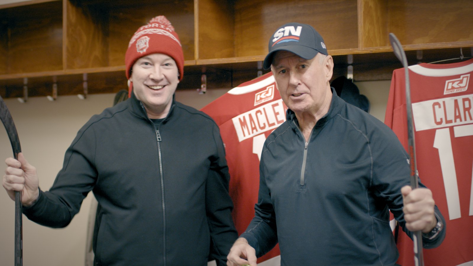 Ron MacLean and Ken Reid in a hockey locker room with stick gloves and team jerseys